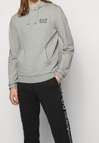 Szara bluza z kapturem z przednią kieszenią, regulowanym sznurkiem oraz logo EA7 na piersi. W zestawie z czarnymi dresami z akcentami logo.