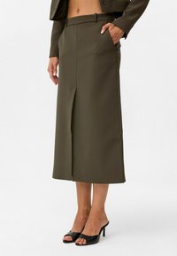 Olijfgroene midi-rok met een voorknop, op maat gemaakte stijl, zijzakken en een gladde textuur, gecombineerd met zwarte open-toe hakken.