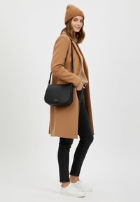 Czarna torebka crossbody z teksturowanego materiału, noszona na jasnobrązowym swetrze, w zestawieniu z beżowym płaszczem i czarnymi dopasowanymi dżinsami, na białym tle.