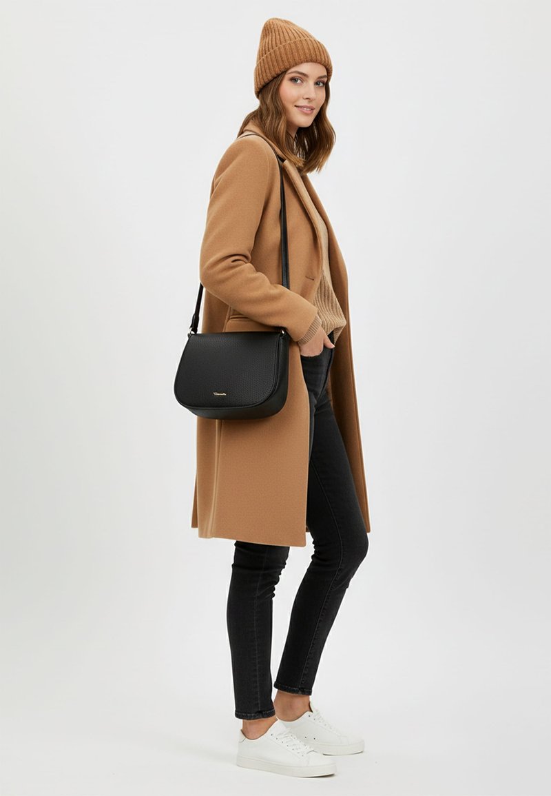 Czarna torebka crossbody z teksturowanego materiału, noszona na jasnobrązowym swetrze, w zestawieniu z beżowym płaszczem i czarnymi dopasowanymi dżinsami, na białym tle.