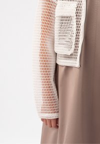 Maglietta bianca a maniche lunghe in rete con motivo a griglia e tessuto testurizzato, dotata di una tasca frontale, abbinata a un indumento liscio di colore beige.