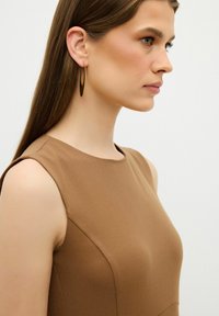 Brun ærmeløs kjole med en glat tekstur, rund halsudskæring og subtile sømme. Guld hoops øreringe bæres, hvilket komplementerer outfittet.