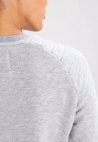 Primo piano della parte superiore della schiena e del collo di una persona che indossa un maglione trapuntato grigio chiaro, di spalle rispetto alla fotocamera.