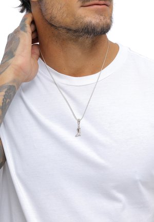 Silberne Kettenhalskette mit einem detaillierten, stiefelförmigen Anhänger, getragen über einem schlichten weißen T-Shirt. Der Hautton ist unter dem Schmuck sichtbar.