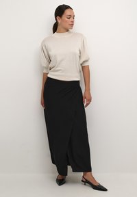 Haut beige à manches courtes avec épaules bouffantes, associé à une jupe portefeuille noire et des escarpins à bride arrière noirs. Textures lisses et lignes épurées.