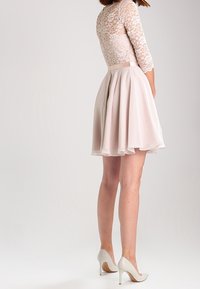 Ljusrosa klänning med spetsliv och trekvartsärmar, har en utsvängd kjol och mjuk textur, parat med vita högklackade skor.