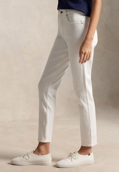 Persoon met witte slim-fit jeans met voorzakken en witte canvas veterschoenen, staand op een neutrale achtergrond.