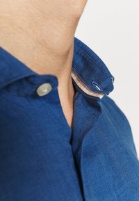 Camicia in lino blu con colletto strutturato, dotata di un sottile dettaglio a righe all'interno del colletto e chiusura con un solo bottone.