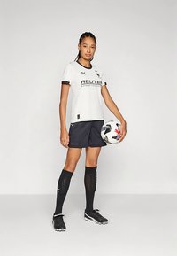 Balta futbolo marškinėliai su juodais elementais, logotipu ir užrašais; derinami su juodais šortais ir juodomis kojinėmis iki kelių. Žaidėjas laiko futbolo kamuolį.