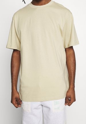 Beiges Baumwoll-T-Shirt mit Rundhalsausschnitt, kurzen Ärmeln und weiter Passform, vor einem schlichten hellen Hintergrund präsentiert.