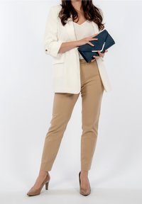 Weißer Blazer, beiger, taillierter Hose und nudefarbene High Heels. Hält eine dunkelblaue Clutch mit silbernem Akzent. Glattes Material und strukturierte Passform.