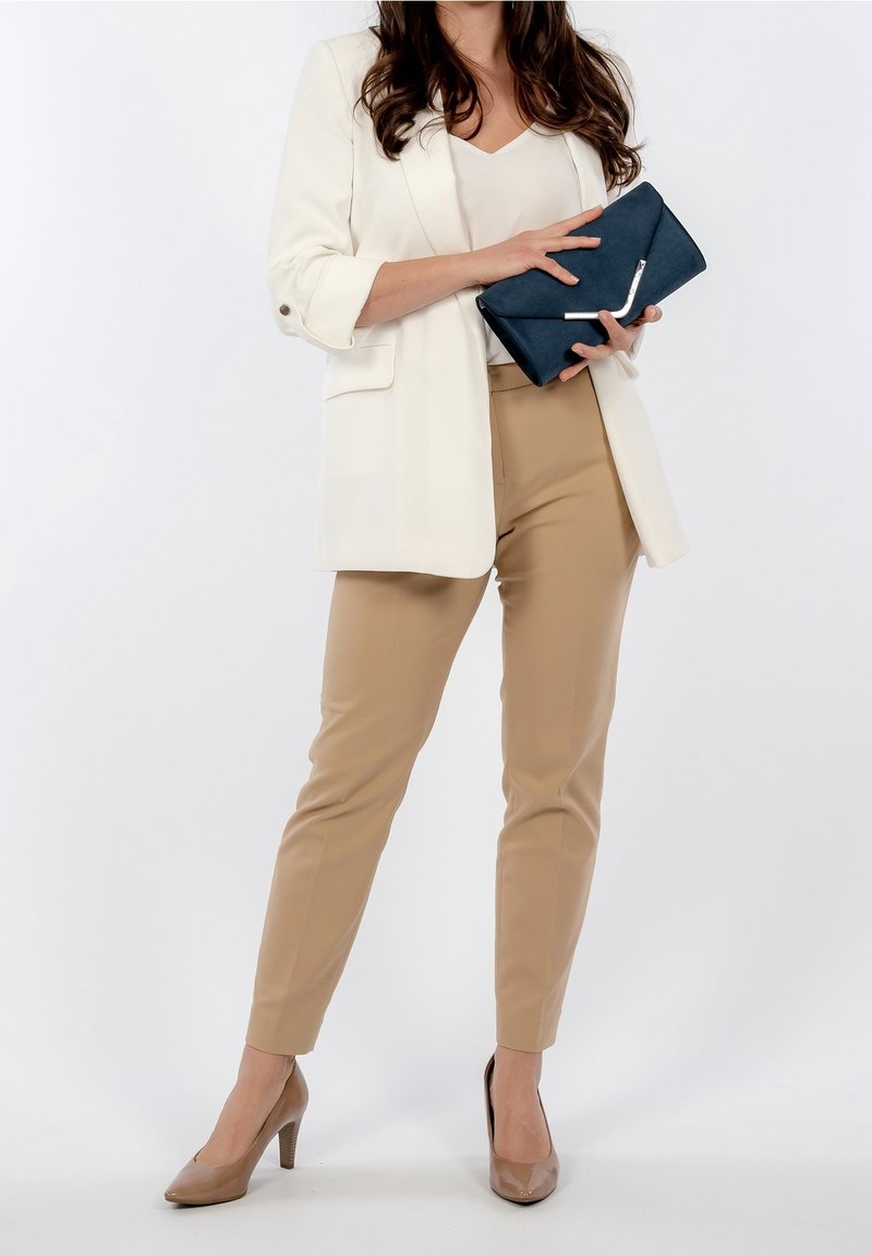 Weißer Blazer, beiger, taillierter Hose und nudefarbene High Heels. Hält eine dunkelblaue Clutch mit silbernem Akzent. Glattes Material und strukturierte Passform.