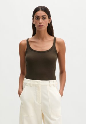 Femme aux longs cheveux foncés portant un débardeur vert foncé et un pantalon blanc taille haute, les mains dans les poches, sur un fond clair.