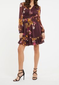 Robe à imprimé floral d'un bourgogne profond avec des manches longues, un col en V, une taille smockée et un ourlet volanté. Associée à des talons noirs à lanières.