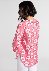 Blouse rose avec motifs floraux blancs, tissu doux, manches longues, bords effilochés et fentes latérales pour plus de détails.