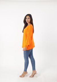 Overmaatse feloranje trui met geribbelde manchet, gecombineerd met strakke blauwe jeans en transparante haksandalen. Effen witte achtergrond.