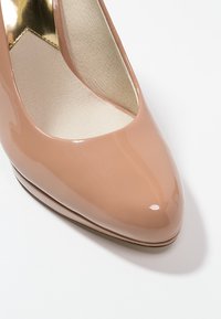 Escarpin en cuir verni beige avec une finition brillante, à bout pointu et un détail découpé à la base du pied. L'intérieur est doublé d'un matériau doux.
