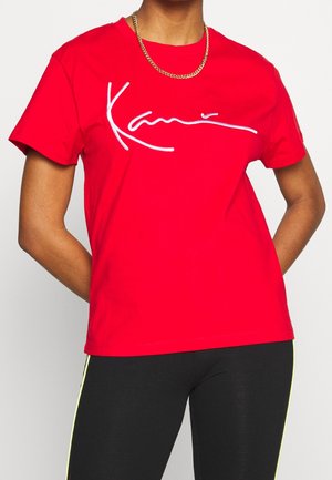 Persoon draagt een felrode T-shirt met witte, handgeschreven tekst over de borst en een gouden ketting, met armen achter de rug.