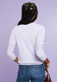 Fialkový top s dlouhým rukávem a kulatým výstřihem, s žebrovanými manžetami a přiléhavým střihem, doplněný modrými džínami a malou burgundskou kabelkou.