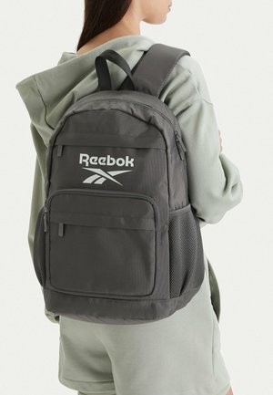 Persona con una mochila Reebok de color gris oscuro, con múltiples compartimentos con cremallera y bolsillos laterales de malla, vestida con sudadera con capucha gris claro y pantalones cortos.