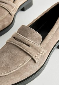 Mocassini in suede color taupe con una finitura liscia. Presentano un accento cucito sulla tomaia e un tacco basso nero. Design pulito e minimalista.