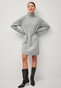 Robe en tricot grise à col haut, avec des poignets et un ourlet côtelés. Associée à des bottes en cuir noir jusqu'aux genoux, mettant en valeur une silhouette ajustée.