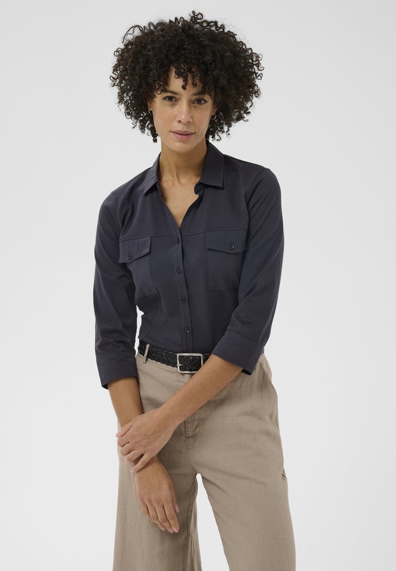 Femme aux cheveux bouclés portant une chemise sombre à boutons avec des poches poitrine et un pantalon beige, debout devant un fond clair uni.