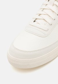 Närbild av tådelen på en vit sneaker med texturerad sula, slätt läderöverdel och vita skosnören mot en ljus bakgrund.