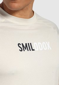Camiseta deportiva de color crema con un patrón texturizado, que presenta la palabra "SMILODOX" en letras negritas negras y blancas en el pecho.
