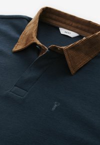 Polo de culoare albastru închis cu un guler texturat din bumbac cord, cu două butoane la gât și un broderie subtilă a logo-ului pe piept.