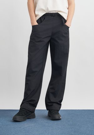 Personne portant un pantalon noir ample avec ceinture réglable et des baskets noires, les mains dans les poches, debout sur un tapis bleu.