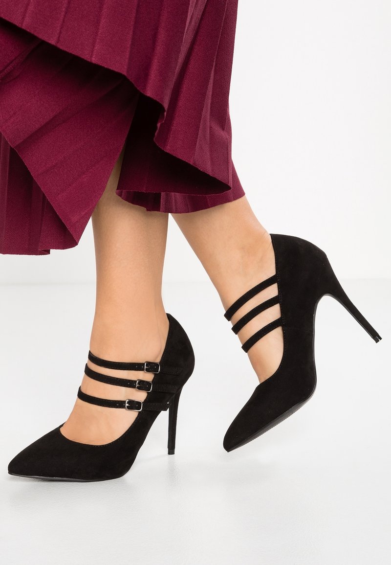 Femme portant des escarpins en daim noir à trois brides avec boucles, assortis à une jupe plissée bordeaux sur un fond blanc.