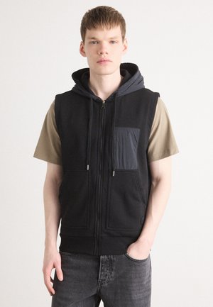Svart ärmlös hoodie med en grå huva och framficka, med dragkedjestängning, stora framfickor och en avslappnad passform, i kombination med en beige skjorta.