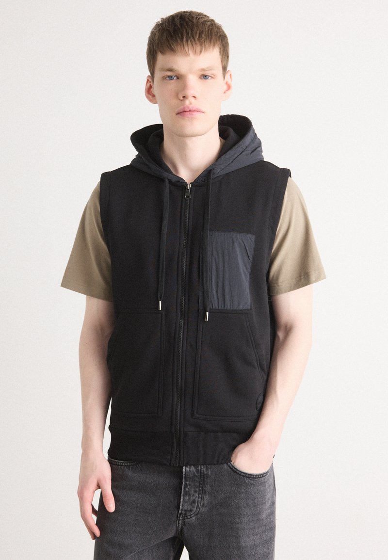 Μαύρο αμάνικο φούτερ με γκρίζα κουκούλα και μπροστινές τσέπες, με κλείσιμο φερμουάρ, μεγάλες μπροστινές τσέπες και χαλαρή εφαρμογή, συνδυασμένο με μπεζ μπλούζα.