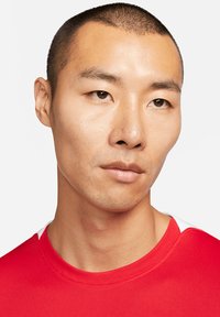 Rotes Sportshirt mit rundem Ausschnitt und weißen Akzenten an den Schultern, getragen von einer Person mit kurzem, eng geschnittenem Haar.