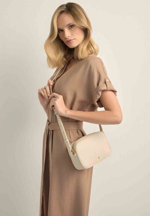 Blondynka w beżowej sukience z paskiem, trzymająca jasnobeżową torebkę na ramię z regulowanym paskiem na jednolitym tle.