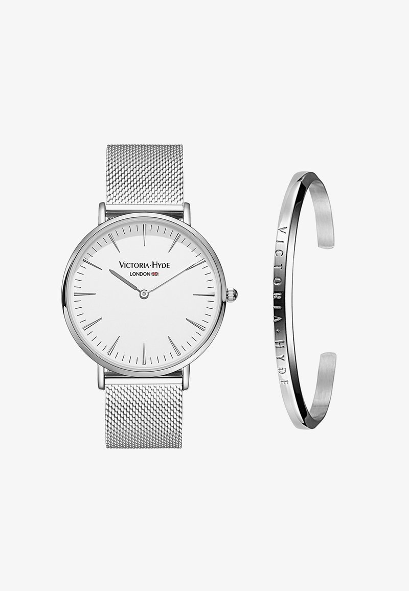 Reloj con correa de malla plateada y esfera blanca, marcadores de horas minimalistas y corona. Incluye una pulsera plateada a juego con un grabado de la marca.