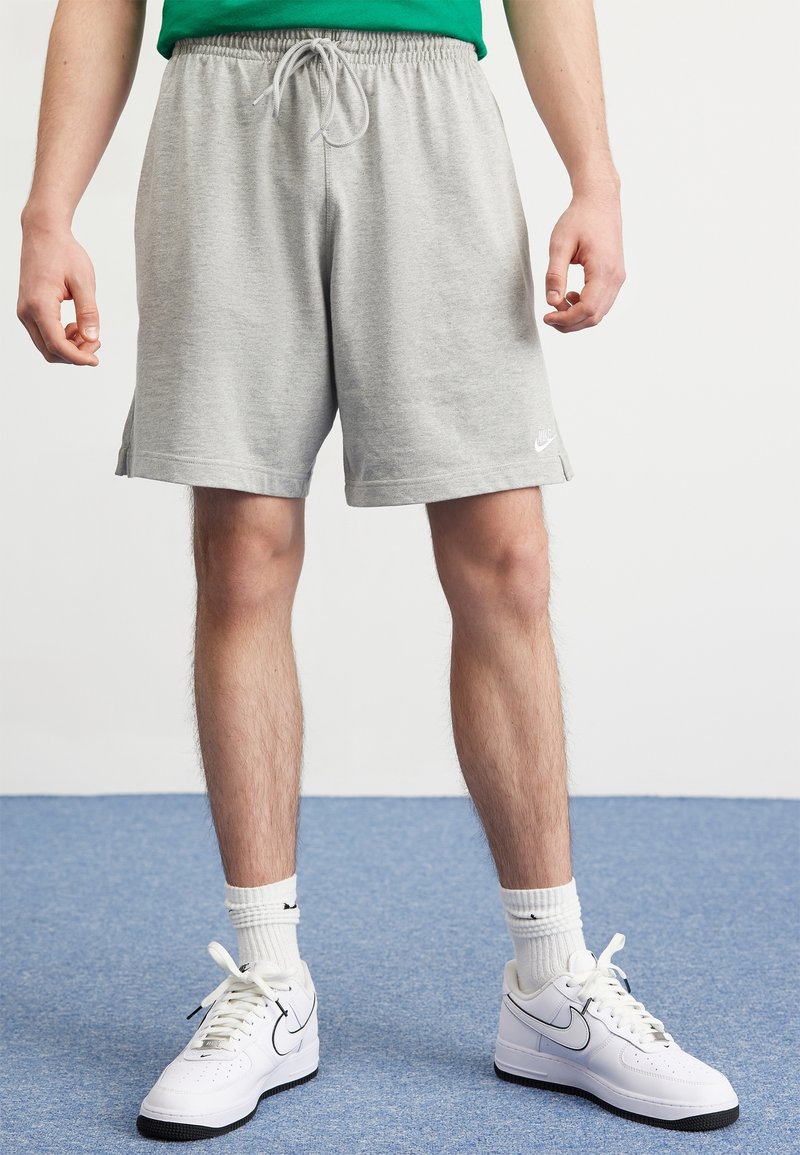 Pantalones cortos deportivos grises con cintura elástica y cordón, con aberturas laterales en el dobladillo y un pequeño logo blanco de Nike. Combinados con zapatillas blancas.