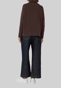 Personne portant un haut marron à manches longues, un jean large foncé et des baskets blanches, se tenant de dos contre un fond uni.