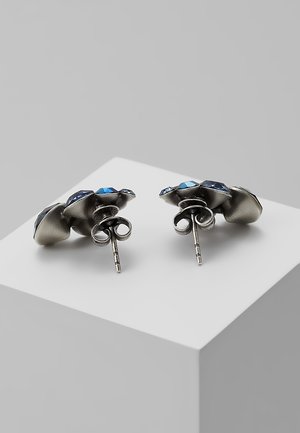 Boucles d'oreilles en argent avec pierres précieuses bleues facettées, vues de dos sur une surface géométrique blanche sur fond gris.