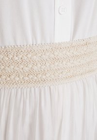 Witte jurk met een geweven crèmekleurige band op de taille. Zachte stof met een gladde afwerking en knoopdetails aan de bovenkant.