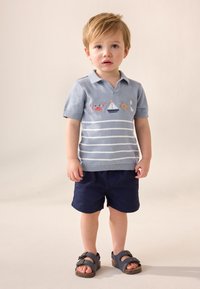 Lyseblå kortærmet polo med hvide striber og nautisk broderi, kombineret med marineblå shorts og grå sandaler. Glat tekstur.