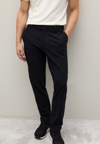 Pantaloni slim fit neri con una texture liscia, dotati di tasche laterali e chiusura con bottone, abbinati a una t-shirt di colore chiaro e sneakers nere.