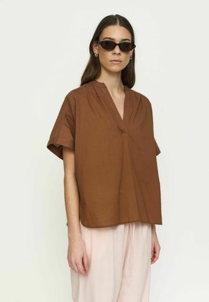 Femme portant un haut marron à manches courtes avec col en V et coupe ample, pantalon beige clair, petits anneaux d'oreilles, et lunettes de soleil ovales noires sur fond uni.