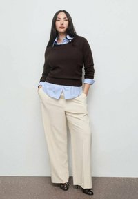 La mujer está de pie con las manos en los bolsillos, vistiendo un suéter marrón oscuro sobre una camisa azul clara, pantalones anchos color crema y tacones abiertos negros.