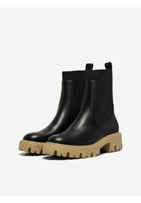 Botas de tobillo de cuero negro con paneles laterales elásticos, suela gruesa en tono marrón claro y un tirador, que presentan una textura suave y una construcción resistente.