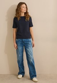 Haut court texturé bleu marine associé à un jean décontracté bleu clair et des baskets blanches, sur fond neutre.