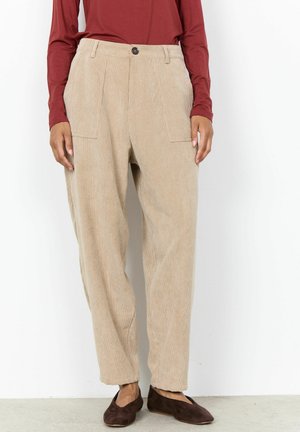 Personne portant un pantalon en velours côtelé beige, des chaussures marron foncé sans lacets et un haut à manches longues couleur rouille, debout sur fond blanc.