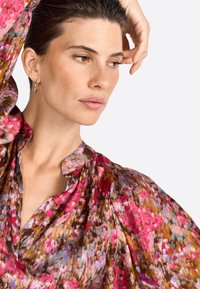 Una donna con capelli scuri posa con aria pensierosa, indossando una blusa floreale colorata con maniche a sbuffo, sullo sfondo di un fondo uniforme.