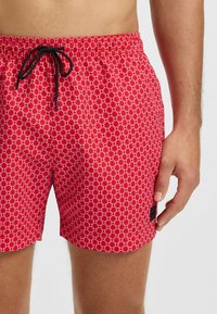 Rode zwemshorts met een wit bloemenpatroon, elastische tailleband met een zwarte trekkoord, en een klein zwart logotag aan de zijkant.